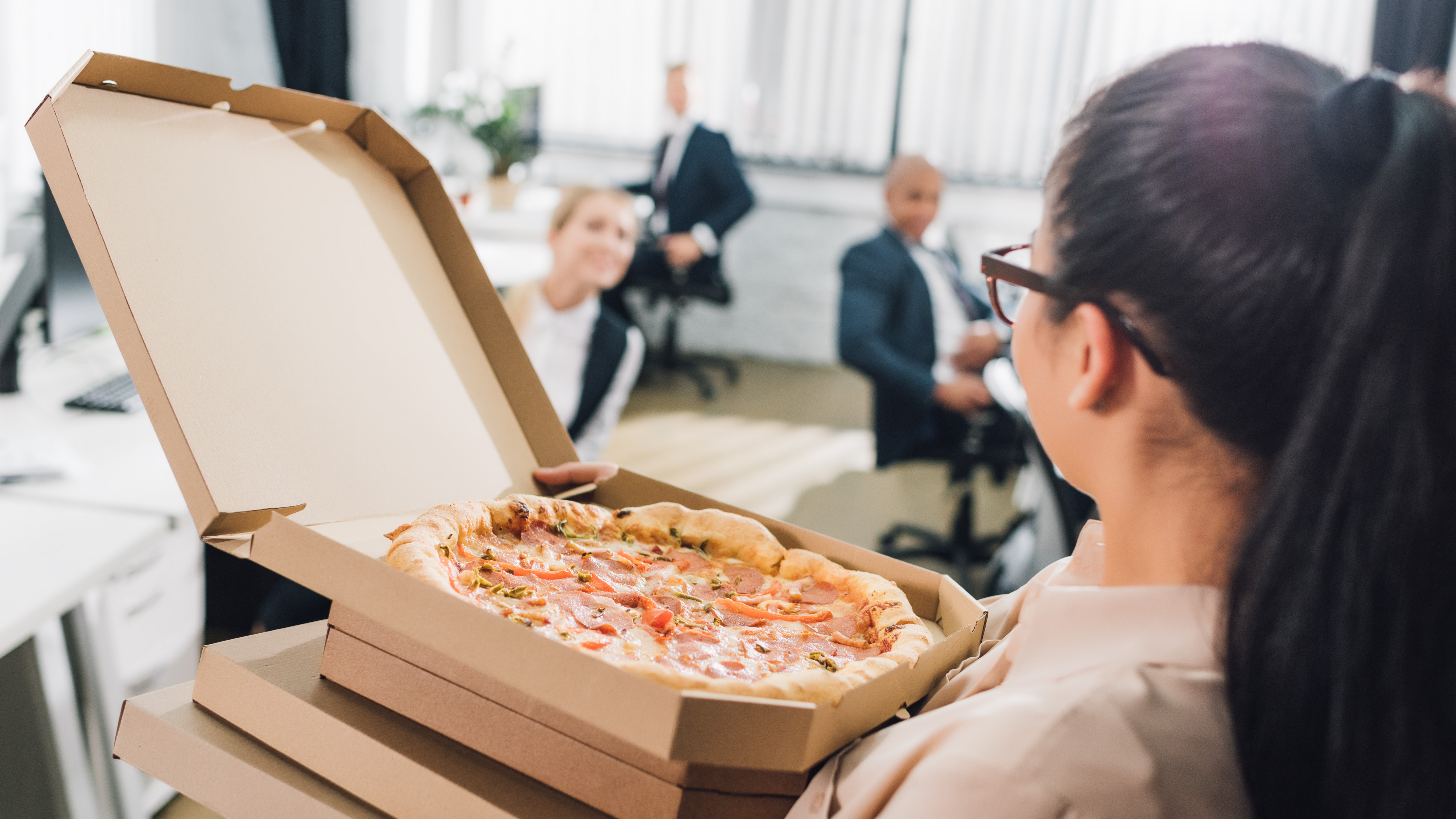 Young woman holding pizza boxes and looking at colleagues in open space office